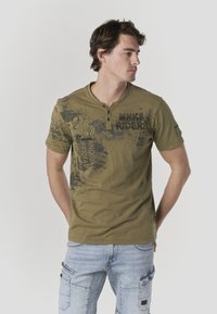 Maglietta henley a maniche corte verde oliva con grafica stampata e placca del colletto; indossata con jeans azzurri chiari con tasche cargo.