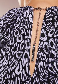 Tissu à motifs noir et blanc avec un décolleté en trou de serrure, présentant un détail froncé et une étiquette en métal avec "Michael Kors" attachée par une chaîne.