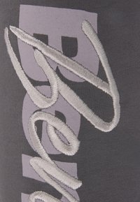 Grauer Stoff mit lila und silberner Stickerei, die abstrakte Textelemente zeigt. Glatte Textur mit geschichteten Designdetails.