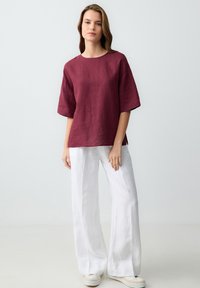 Top in lino burgundy con maniche corte, scollatura tonda e spacchi laterali, abbinato a pantaloni in lino bianchi a gamba larga e sneakers casual.