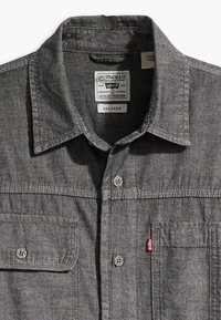 Camicia grigia con bottoni realizzata in un tessuto con una leggera texture, dotata di colletto, due tasche frontali e una linguetta rossa Levi's sulla cucitura laterale.