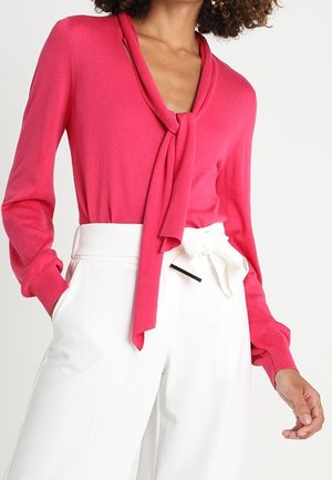 Kobieta ubrana w jasnoróżową bluzkę z długim rękawem, z wiązaniem przy dekolcie, oraz białe spodnie z wysokim stanem z detalem w formie kokardy na talii.