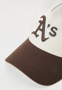 Kasket med en beige krone og brun skygge, der har et stort broderet "A's" i brun og lys blå. tekstureret stof med syning detaljer.