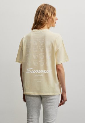 Pitkätukkainen nainen, jolla on beige T-paita valkoisin juomalasin kuvin ja teksti "Summer Mode On", yhdistettynä vaaleanharmaisiin housuihin.