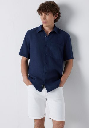 Junger Mann mit lockigem Haar, der ein dunkelblaues kurzärmeliges Hemd mit Knöpfen und weiße hochgekrempelte Shorts trägt, steht an einer weißen Wand.