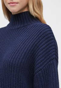 Jūras zila adīta džemperis ar augstu ribotu apkakli, kas izceļ fakturētu audumu un platas piedurknes. Tuva bilde uz kakla un plecu apvidu.