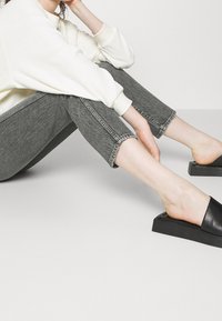 Šedé zkrácené džíny s roztrženým lemem a hladkým svetrem v barvě slonové kosti. Černé slip-on boty mají robustní podrážku.