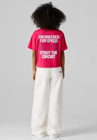 Osoba s kučeravými vlasmi oblečená v ružovom krátkom tričku s mapou pretekárskeho okruhu a bielych nohaviciach s rozšírenými nohavicami, stojaca pred sivým pozadím.