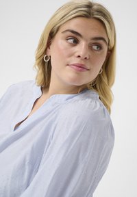 Blusa azzurro chiaro con sottili righe verticali e piccoli punti bianchi, caratterizzata da un collo a V e un tessuto morbido e fluido.