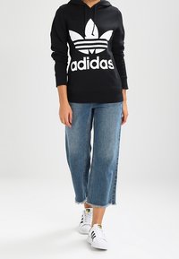 Person som bär en svart Adidas-hoodie, avklippta blå jeans och vita Adidas-sneakers med svarta ränder och går framåt.