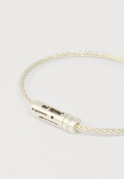Sudraba savītas kabeli aproce ar cilindrisku sprādzi, uz kuras ir iegravēts "le gramme" un mehāniskas detaļas uz gaiša fona.