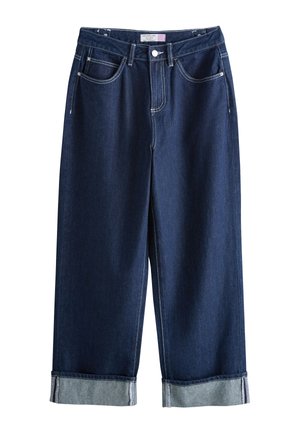 Vrouw staat blootsvoets, draagt een witte top met een vierkante halslijn en donkerblauwe wijde jeans met omslag, handen in de zakken.