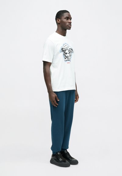 Λευκό κοντομάνικο μπλουζάκι με γραφικό της Μέδουσας, σε συνδυασμό με χαλαρά τιρκουάζ παντελόνια και μαύρα αθλητικά παπούτσια. Απλή, καθημερινή εμφάνιση με τολμηρές λεπτομέρειες.