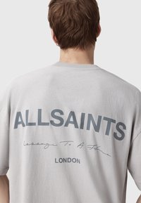 T-shirt en coton gris avec un grand logo "ALLSAINTS" et un texte en script au dos, avec un col rond et une coupe décontractée.