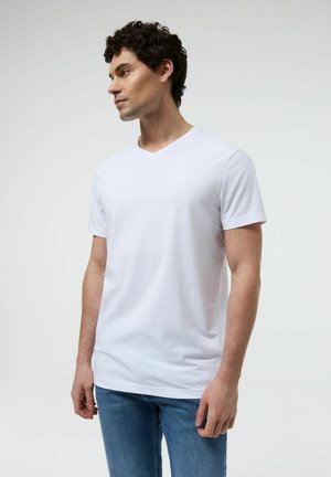 Mężczyzna w białym, gładkim t-shircie z krótkim rękawem i dekoltem w serek oraz niebieskich dżinsach, stoi i patrzy w lewo na białym tle.