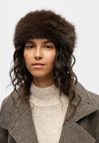 Chapeau en fourrure marron avec un design épais et rond, associé à un pull en tricot crème et un manteau texturé gris, créant un look superposé.