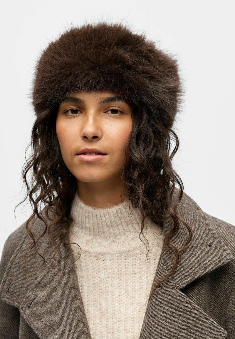 Chapeau en fourrure marron avec un design épais et rond, associé à un pull en tricot crème et un manteau texturé gris, créant un look superposé.