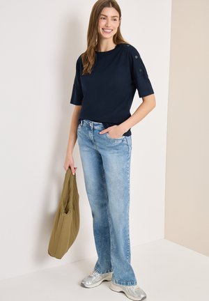 Femme souriante, portant un haut bleu marine à manches courtes avec des boutons, un jean bleu clair, des baskets argentées, tenant un sac vert olive dans un environnement intérieur neutre.