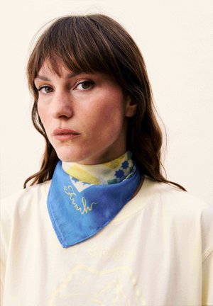 Femme aux cheveux bruns portant une chemise crème et un foulard bleu à motifs autour du cou, sur un fond clair uni.