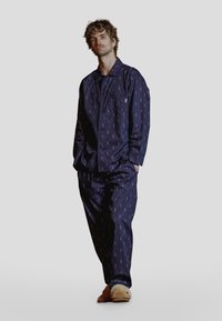 Marineblå pysjamasett i mykt stoff med et subtilt trykk. Inneholder en skjorte med knapper og lomme, samt matchende bukser. Komplettert med beige tøfler.