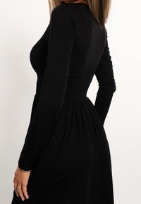 Robe noire à manches longues avec un corsage ajusté et une jupe évasée, en tissu lisse. Elle présente un col rond et une taille froncée.