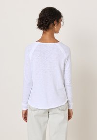 Frau mit lockigem Haar in weißem Langarmshirt und hellen Jeans steht vor einer schlichten, cremeweißen Wand.