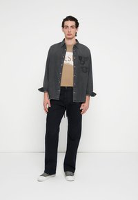 Ciemnoszara koszula dżinsowa na guziki z haftowanymi naszywkami, noszona na beżowym t-shircie. Do tego granatowe spodnie i szaro-białe tenisówki.