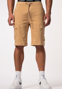 Shorts cargo beiges avec des ourlets retroussés, deux poches latérales et une taille à cordon. Portés avec des baskets blanches et des chaussettes de mi-mollet.