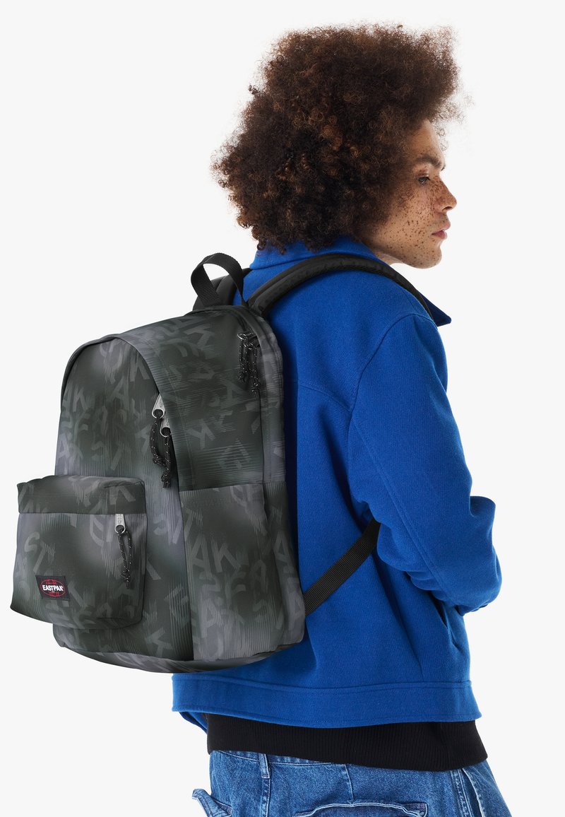 Personne aux cheveux bouclés portant une veste bleue et un sac à dos gris foncé à motifs avec une poche avant et des bretelles noires.