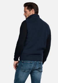 Marineblaues Strickweste mit hohem Kragen und geripptem Stoff, kombiniert mit dunkelblauen Langarmshirts. Dazu getragen wurden hellblaue Denim-Jeans.