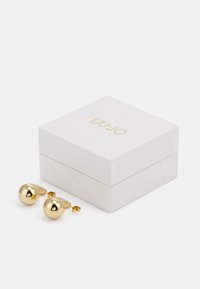 Goldfarbene Ohrstecker mit einem glatten, runden Design, neben einer weißen Box mit goldener Beschriftung "LIU JO".