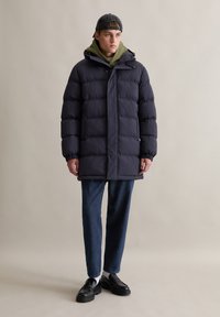 Dunkelblauer Puffy-Mantel mit Kapuze, ausgestattet mit einem gesteppten Design, elastischen Bündchen und einem Knopfverschluss, kombiniert mit dunklen Jeans und schwarzen Schuhen.