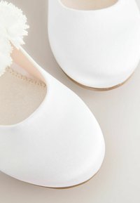 White fabric shoes with a smooth texture, round toe design, and a flower accent on the side. Flat soles with a light brown edge.