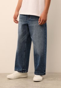 Hele sinised, laia lõikega teksapüksid tuhmunud viimistlusega, ülespööratud äärega ja standardses viie taskuga disainis, kombineeritud valgete tossudega.