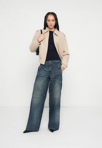 Casaco bomber bege cropped com gola, top canelado azul-marinho e jeans de ganga azul de perna larga com costuras inclinadas e saltos escuros.