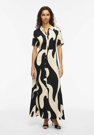 Kobieta w długiej sukience na guziki z abstrakcyjnym wzorem w kolorze czarno-beżowym, z krótkim rękawem i czarnych butach na obcasie z odkrytymi palcami, stoi na białym tle.