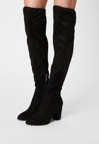 Černé semišové kozačky nad kolena s špičatou špičkou a robustním blokovým podpatkem. Mají boční zip a hladký povrch.