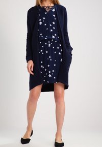 Kvinna iklädd en marinblå klänning med stjärnmönster, tillsammans med en matchande lång ribbad cardigan och svarta lågklackade skor, stående med händerna i fickorna.