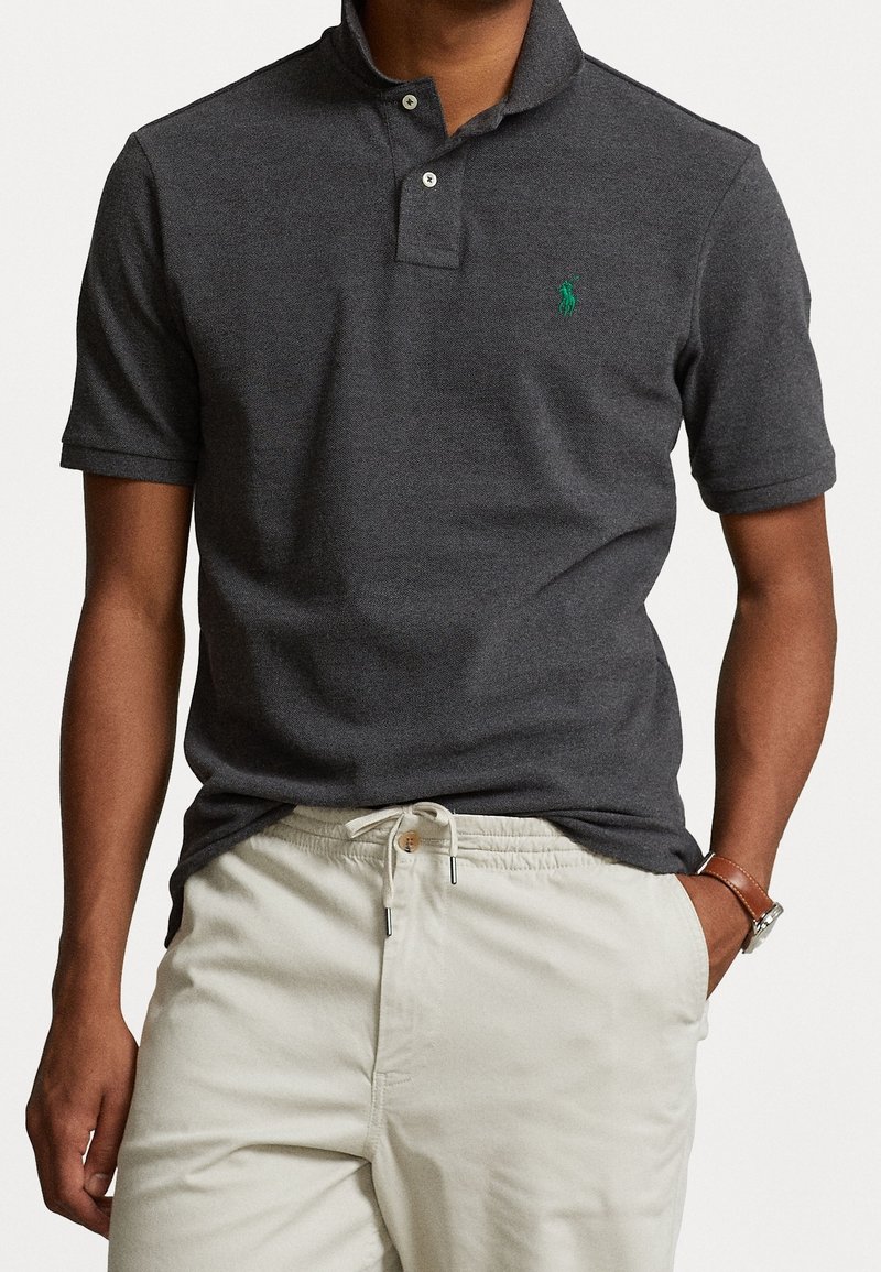 Homme portant un polo gris foncé à manches courtes avec un logo vert et un pantalon beige clair avec cordon, main gauche dans la poche, sur fond blanc.