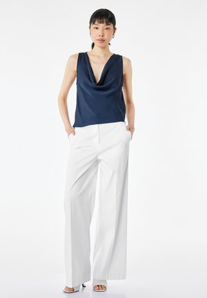Mujer con pelo negro de pie, lleva una blusa sin mangas azul marino con drapeado, pantalones anchos blancos y tacones abiertos blancos, manos en los bolsillos.