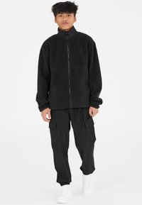 Svart fleecjacka med hög krage och framdragkedja, kombinerad med svarta cargobyxor som har sidofickor och elastiska muddar. Vita sneakers.