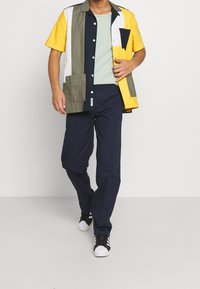 Mångfärgad kortärmad skjorta med gula, gröna, vita och marinblå paneler, tillsammans med marinblå byxor och svarta sneakers.