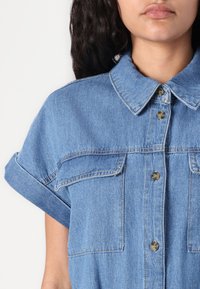 Svetlo modra denim srajca s kratkimi zvitimi rokavi, ovratnikom in dvema žepoma na prsih. Ima kovinske gumbe in mehak otip.