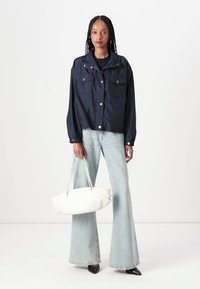 Giacca cropped blu navale con due tasche frontali, abbinata a jeans svasati di un blu chiaro e una borsa bianca. Modella che indossa scarpe nere a punta.