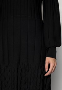 Abito nero in maglia con texture a coste verticali nella parte superiore e un motivo ondulato in rilievo all'orlo. Maniche lunghe a palloncino e polsini aderenti.