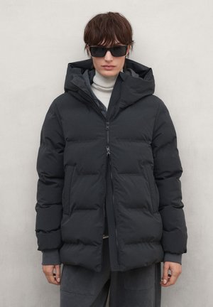 Persona con chaqueta negra acolchada con capucha, cuello alto gris debajo, pantalones oscuros y gafas de sol negras, de pie frente a un fondo claro y liso.