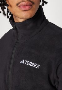 Svart fleecejakke med høy krage, glidelås og hvit "TERREX" logo på brystet. Myk tekstur, ingen synlige mønstre eller aksenter.