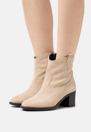 Botines de ante beige con punta afilada y tacón en bloque, con costuras decorativas y una pestaña en la parte posterior.