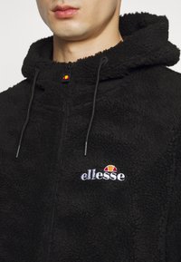 Svart fleece hettegenser med glidelås, med teksturert stoff, hette og en brodert "ellesse"-logo i hvitt, rødt og gult.
