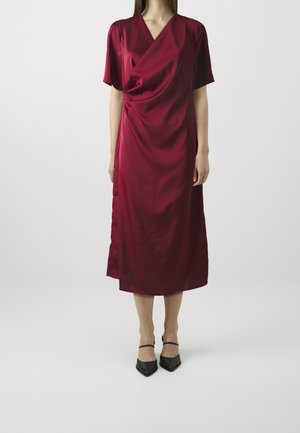 Kobieta ubrana w sukienkę z czerwonego, satynowego materiału o długości do kolan, z krótkimi rękawami i marszczonym przodem, nosząca czarne szpilki z paskami wokół kostek.